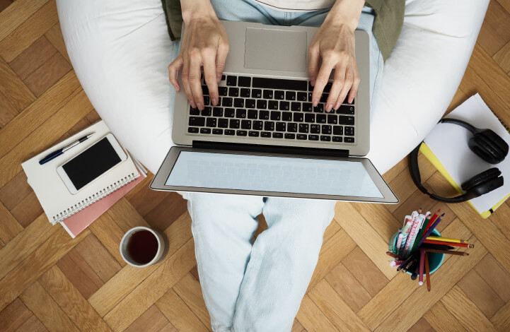 An employee is seen working on his laptop in an informal work setup, seated on a beanbag with a coffee mug, notes and headphones around him